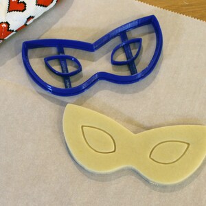 May include: A blue plastic cookie cutter in the shape of a masquerade mask sits next to a cookie cut in the same shape. The cookie is light yellow and has the eye shapes imprinted on it. A red and white heart-patterned oven mitt is in the background.