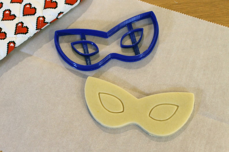 May include: A blue plastic cookie cutter in the shape of a masquerade mask sits next to a cookie cut in the same shape. The cookie is light yellow and has the eye shapes imprinted on it. A red and white heart-patterned oven mitt is in the background.