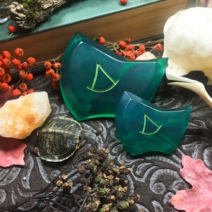 May include: Two green resin dice with a stylized letter "D" on each face. The dice are on a dark brown surface with a skull, a turtle, and a small pile of dried plants.