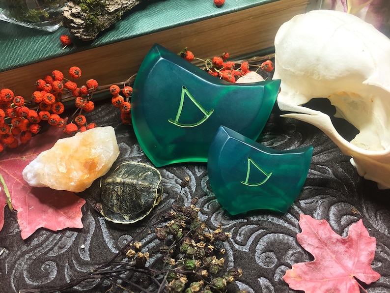 May include: Two green resin dice with a stylized letter "D" on each face. The dice are on a dark brown surface with a skull, a turtle, and a small pile of dried plants.