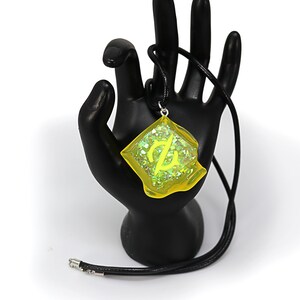 May include: A yellow resin pendant necklace with a green glitter embedded inside. The pendant features a stylized symbol and is hanging from a black cord.