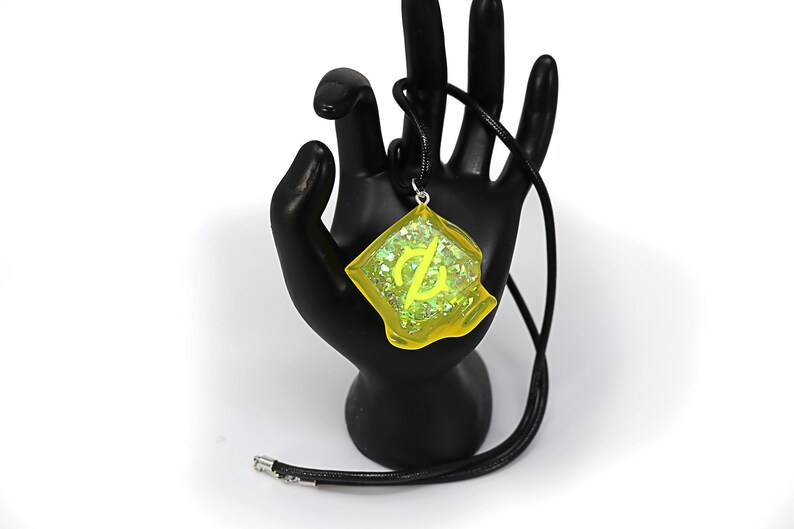 May include: A yellow resin pendant necklace with a green glitter embedded inside. The pendant features a stylized symbol and is hanging from a black cord.