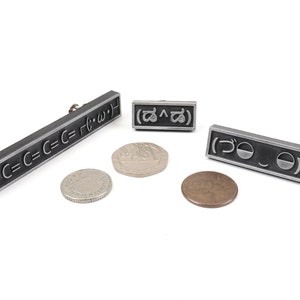 May include: Three silver metal pins with different symbols. The first pin has the text "C=C=C=C=C= r(&bull;&omega;&bull;)". The second pin has the text "(^)". The third pin has the text "づ".