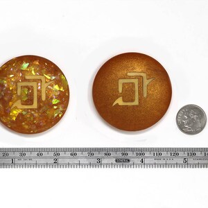 May include: Two round, gold-colored, decorative coasters with a stylized symbol in the center. The symbol is a stylized "5" with a small arrow pointing to the right. One coaster has a glittery, iridescent finish, while the other has a smooth, metallic finish.