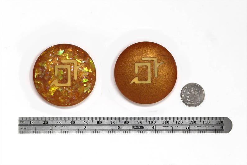 May include: Two round, gold-colored, decorative coasters with a stylized symbol in the center. The symbol is a stylized "5" with a small arrow pointing to the right. One coaster has a glittery, iridescent finish, while the other has a smooth, metallic finish.