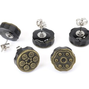 以下が含まれることがあります： 拳銃のシリンダーデザインのスタッドピアス5つ。ピアスは黒いベースに、ブロンズ色の詳細なトップが付いています。ポストはシルバーカラーです。ピアスは白い背景に表示されています。