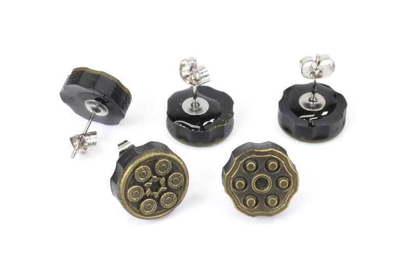 以下が含まれることがあります： 拳銃のシリンダーデザインのスタッドピアス5つ。ピアスは黒いベースに、ブロンズ色の詳細なトップが付いています。ポストはシルバーカラーです。ピアスは白い背景に表示されています。