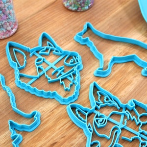 May include: Four blue plastic dog cookie cutters. Two are of dog heads, and two are of dogs standing on all fours.