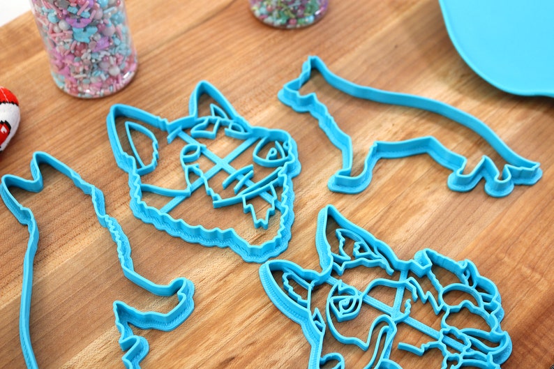 May include: Four blue plastic dog cookie cutters. Two are of dog heads, and two are of dogs standing on all fours.
