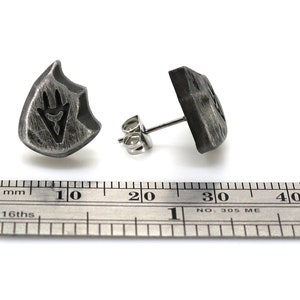 Puede incluir: Un par de pendientes de plata con un dise&ntilde;o en forma de escudo. El escudo tiene un s&iacute;mbolo estilizado en su interior. Los pendientes se muestran junto a una regla, midiendo aproximadamente 1,5 cm de di&aacute;metro.