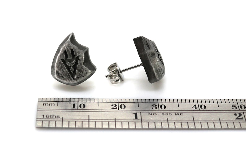 Puede incluir: Un par de pendientes de plata con un dise&ntilde;o en forma de escudo. El escudo tiene un s&iacute;mbolo estilizado en su interior. Los pendientes se muestran junto a una regla, midiendo aproximadamente 1,5 cm de di&aacute;metro.