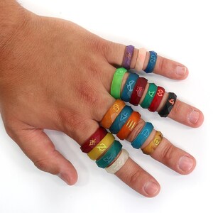 May include: A hand wearing a variety of colorful silicone rings with different symbols and designs. The rings are stacked on each finger.