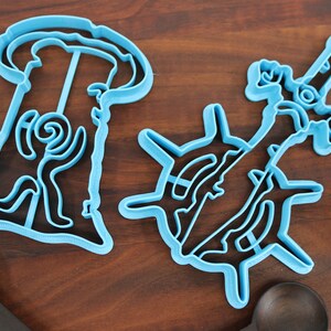 Puede incluir: Dos cortadores de galletas de pl&aacute;stico azul, uno con forma de persona y el otro con forma de espada.