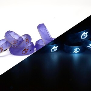 May include: A set of six purple glow-in-the-dark rings with a red flame design. The rings are stacked on a white surface and glow brightly in the dark.