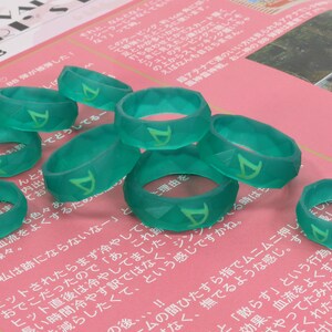 Pu&ograve; includere: Un gruppo di otto anelli in silicone verde con un design geometrico e una lettera 'D' stilizzata su ogni anello.