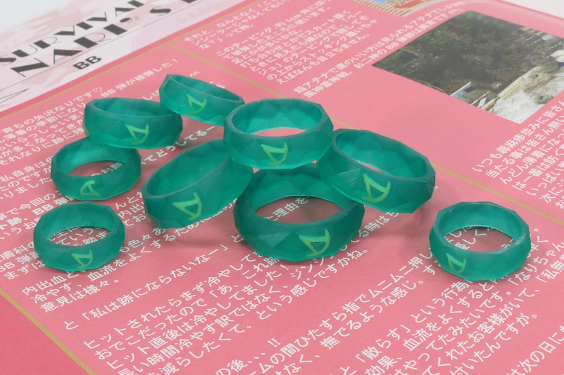 Pu&ograve; includere: Un gruppo di otto anelli in silicone verde con un design geometrico e una lettera 'D' stilizzata su ogni anello.
