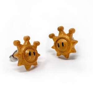 May include: A pair of gold-coloured earrings shaped like a sun with a face. The earrings are made of metal and have a shiny finish.