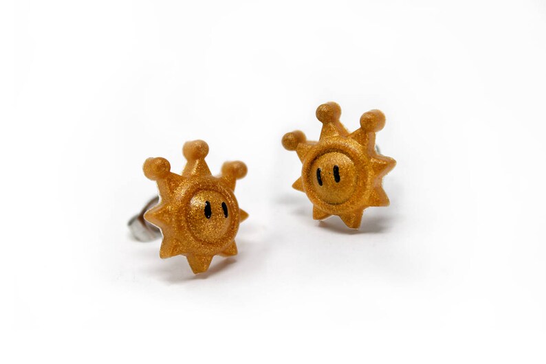 May include: A pair of gold-coloured earrings shaped like a sun with a face. The earrings are made of metal and have a shiny finish.