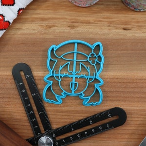 May include: Blue plastic cookie cutter shaped like a cartoon character with a flower in their hair. The character has a wide smile and their tongue is sticking out.