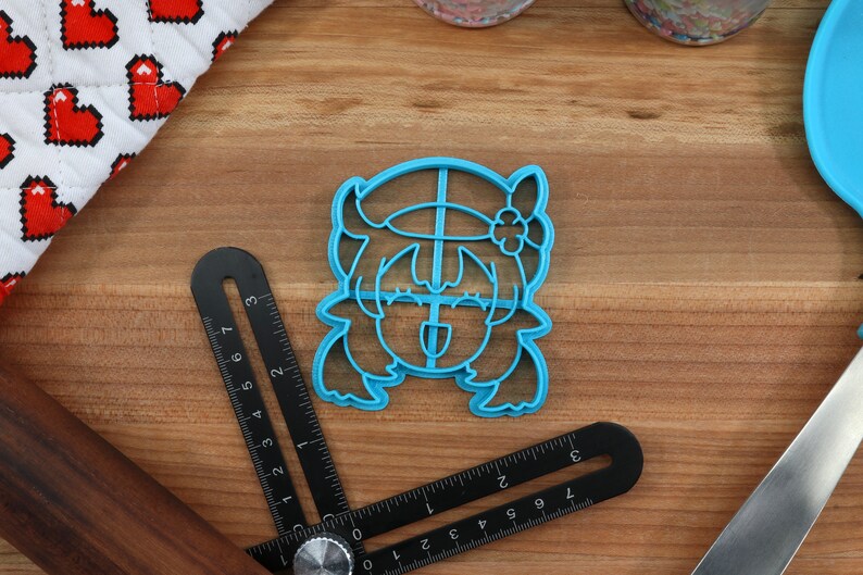 May include: Blue plastic cookie cutter shaped like a cartoon character with a flower in their hair. The character has a wide smile and their tongue is sticking out.