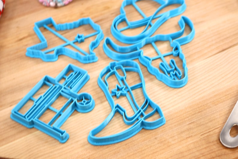 May include: A set of five blue plastic cookie cutters shaped like a cowboy boot, a cowboy hat, a longhorn skull, a wagon, and the state of Texas.