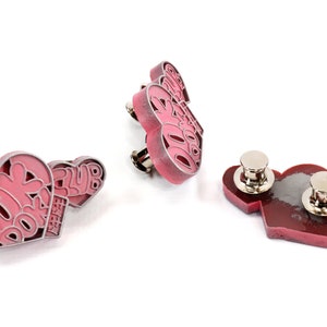 May include: Three pink heart-shaped pins with the text "DORK DORK" and "CLUB EST. 2018" on them. The pins have silver metal backs with a safety pin closure.