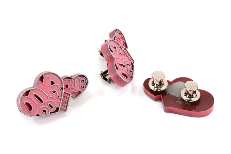 May include: Three pink heart-shaped pins with the text "DORK DORK" and "CLUB EST. 2018" on them. The pins have silver metal backs with a safety pin closure.