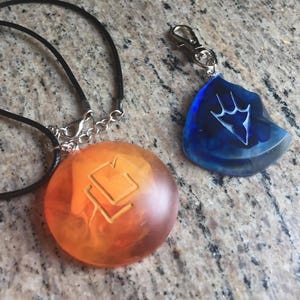 May include: Two necklaces with pendants featuring different symbols. One pendant is a round, orange-colored glass with a stylized symbol. The other pendant is a blue, teardrop-shaped glass with a stylized symbol.