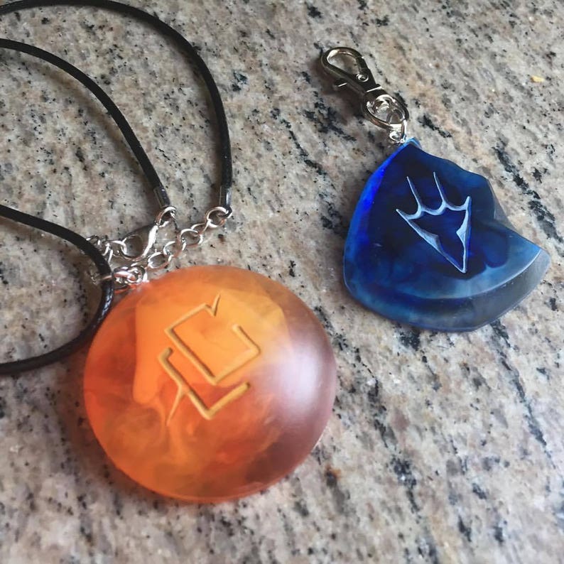 May include: Two necklaces with pendants featuring different symbols. One pendant is a round, orange-colored glass with a stylized symbol. The other pendant is a blue, teardrop-shaped glass with a stylized symbol.