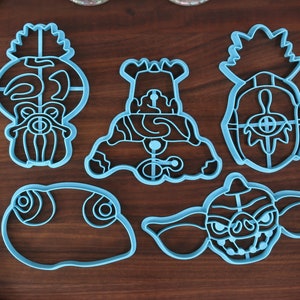May include: Five blue plastic cookie cutters shaped like different characters from the movie Gremlins. The cutters are on a wooden surface.