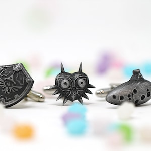 May include: Three silver cufflinks featuring iconic symbols from the Legend of Zelda video game series: the Hylian Shield, the Majora's Mask, and the Ocarina of Time.