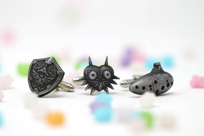 May include: Three silver cufflinks featuring iconic symbols from the Legend of Zelda video game series: the Hylian Shield, the Majora's Mask, and the Ocarina of Time.