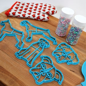 May include: A set of blue plastic dog-shaped cookie cutters, a rolling pin, a spatula, and two jars of colorful sprinkles on a wooden cutting board.