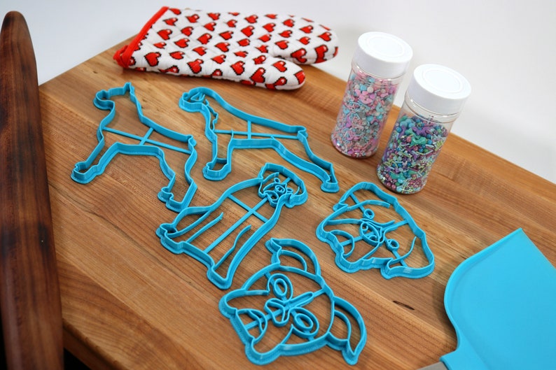 May include: A set of blue plastic dog-shaped cookie cutters, a rolling pin, a spatula, and two jars of colorful sprinkles on a wooden cutting board.