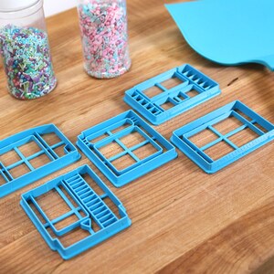 May include: Five blue plastic cookie cutters shaped like windows, on a wooden surface. Two jars of sprinkles are in the background.
