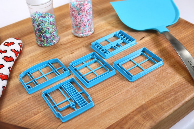 May include: Five blue plastic cookie cutters shaped like windows, on a wooden surface. Two jars of sprinkles are in the background.
