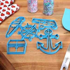 May include: A collection of blue plastic nautical-themed cookie cutters, including an anchor, ship's wheel, and treasure chest. Also present are a blue spatula, a heart-patterned oven mitt, a rolling pin, and jars of sprinkles, all arranged on a wooden surface.