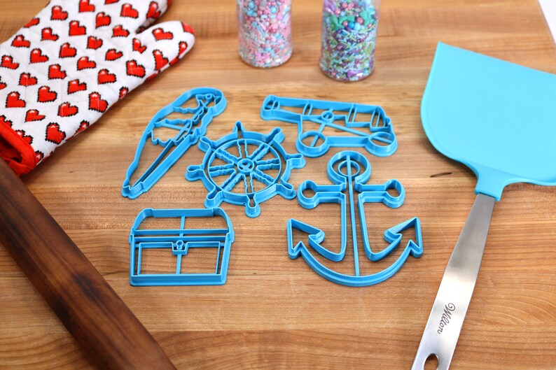May include: A collection of blue plastic nautical-themed cookie cutters, including an anchor, ship's wheel, and treasure chest. Also present are a blue spatula, a heart-patterned oven mitt, a rolling pin, and jars of sprinkles, all arranged on a wooden surface.