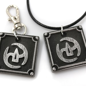 May include: Two silver-coloured pendants with a black and silver symbol on a black background. One pendant is on a black cord necklace and the other is on a silver key chain.