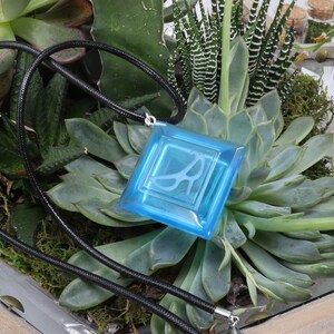 May include: A blue glass pendant with a white design hangs from a black cord. The pendant is displayed in a terrarium with green succulents.