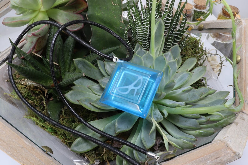 May include: A blue glass pendant with a white design hangs from a black cord. The pendant is displayed in a terrarium with green succulents.