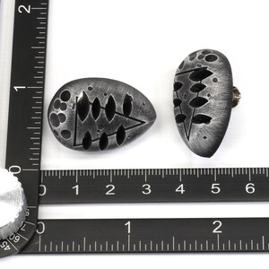 May include: Two silver-colored, oval-shaped, decorative knobs with a black and white design. The knobs have a textured surface and a small screw on the back.