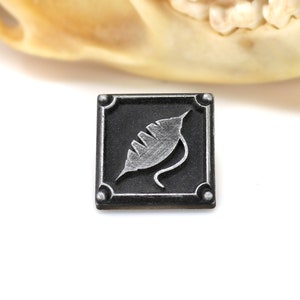 May include: A small, square, black metal tile with a silver leaf design. The tile has a raised border and four silver studs.