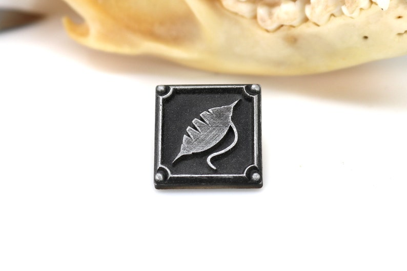 May include: A small, square, black metal tile with a silver leaf design. The tile has a raised border and four silver studs.