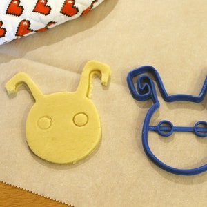 May include: A blue plastic cookie cutter shaped like a cartoon character with large ears and two eyes. Next to it is a yellow cookie that has been cut out using the cookie cutter.
