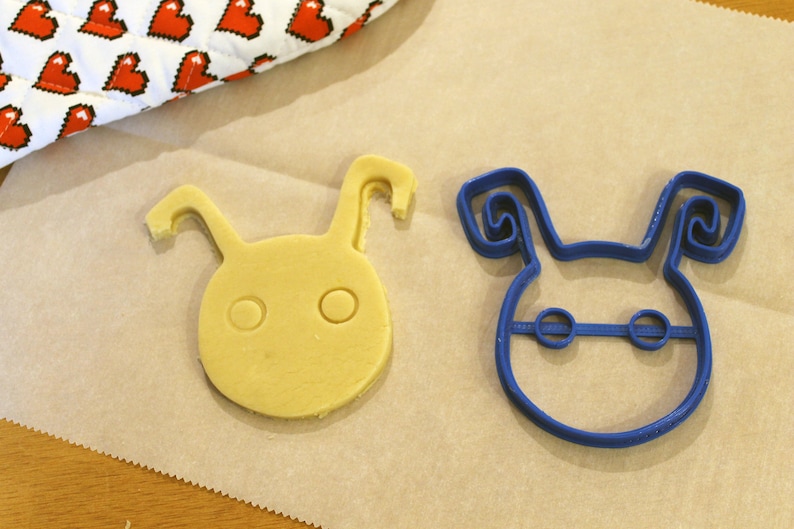 May include: A blue plastic cookie cutter shaped like a cartoon character with large ears and two eyes. Next to it is a yellow cookie that has been cut out using the cookie cutter.