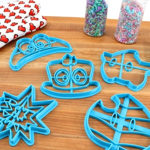 May include: A set of six blue plastic cookie cutters shaped like a hat, a cake, a spaceship, a snowman, a star, and a leaf. The cutters are on a wooden surface.