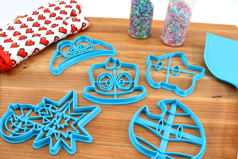 May include: A set of six blue plastic cookie cutters shaped like a hat, a cake, a spaceship, a snowman, a star, and a leaf. The cutters are on a wooden surface.