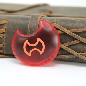May include: A red, crescent-shaped pendant with a stylized orange symbol. The pendant is made of glass and has a smooth, glossy finish.