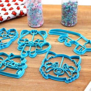 May include: Five blue plastic cookie cutters shaped like red pandas. The cutters are on a wooden surface with two jars of sprinkles in the background.